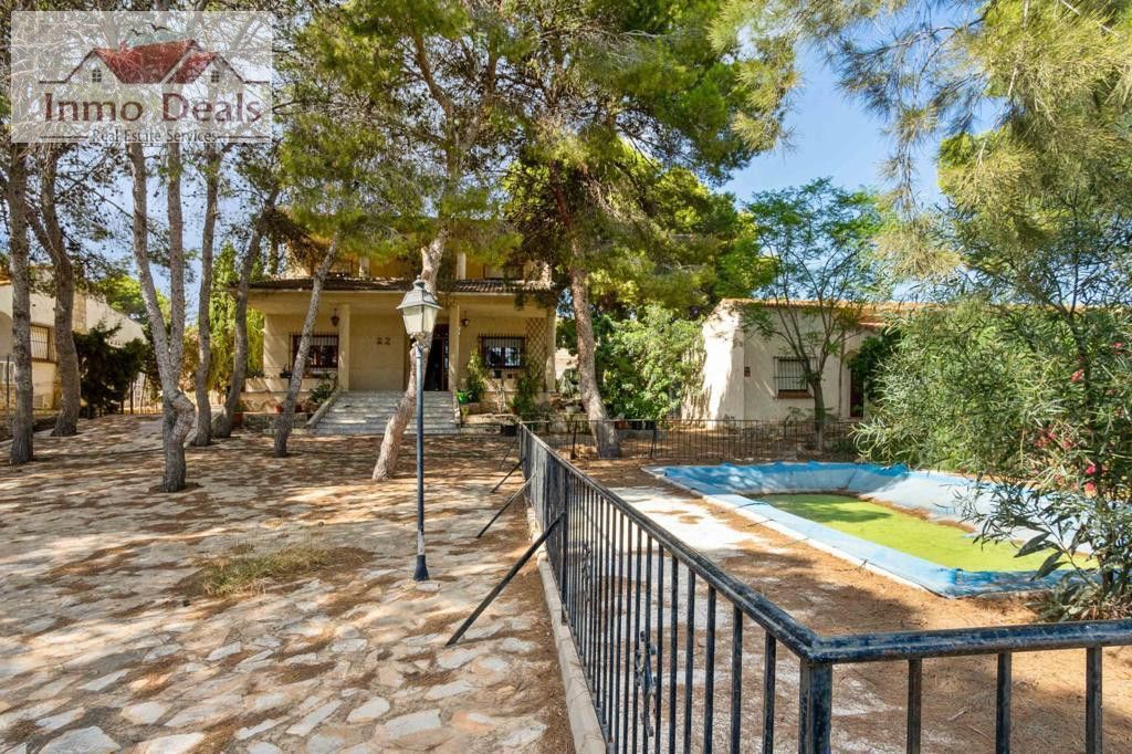 Villa med trädgård, träd, pool och uthus under solig himmel.